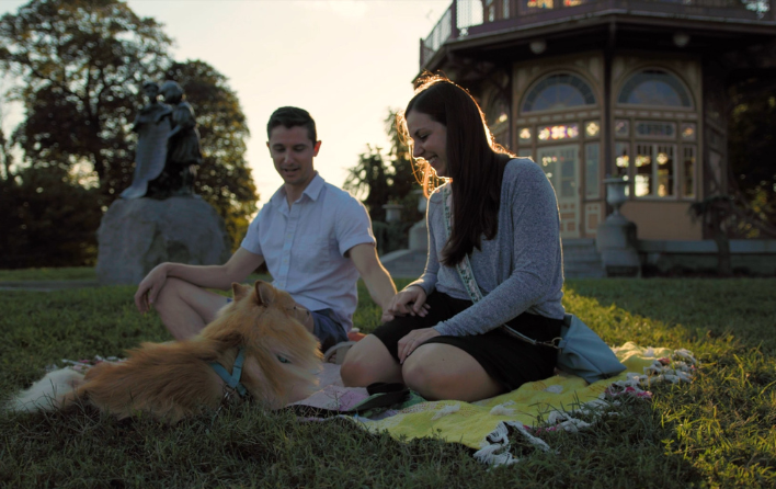 Two people sit outside on a blanket on a lawn at dusk with a small dog.
